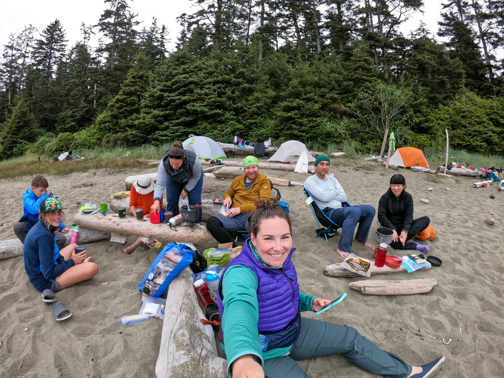 A group of backpackers cooking on the beach while a hiker in a MEC Boundary Light vest takes a selfie