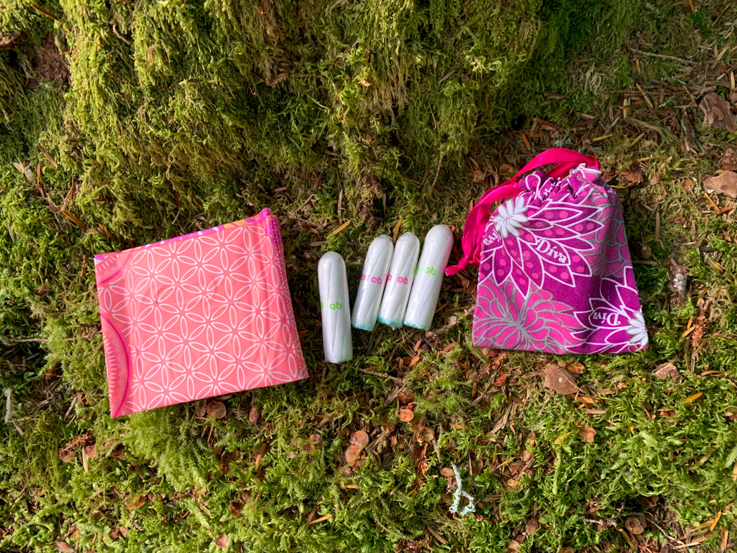 Menstrual products including pads, tampons and a cup rest on a mossy forest floor. Learn how to camp and hike on your period