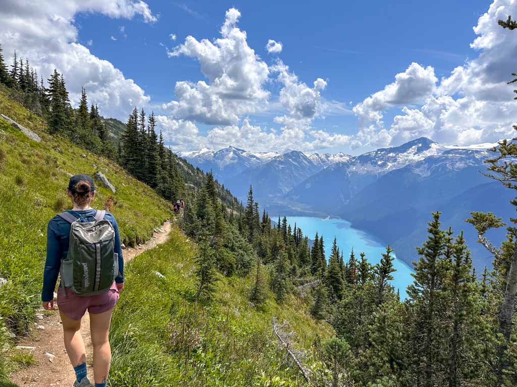 A hiker with a black backpack hikes along the High Note Trail with Cheakamus Lake in the background.