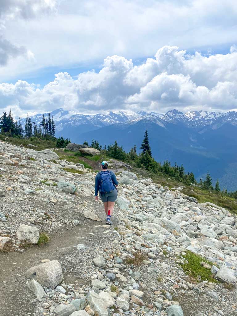 A female hiker wearing a blue backpack walks along a rocky trail with snow capped mountains in the background.