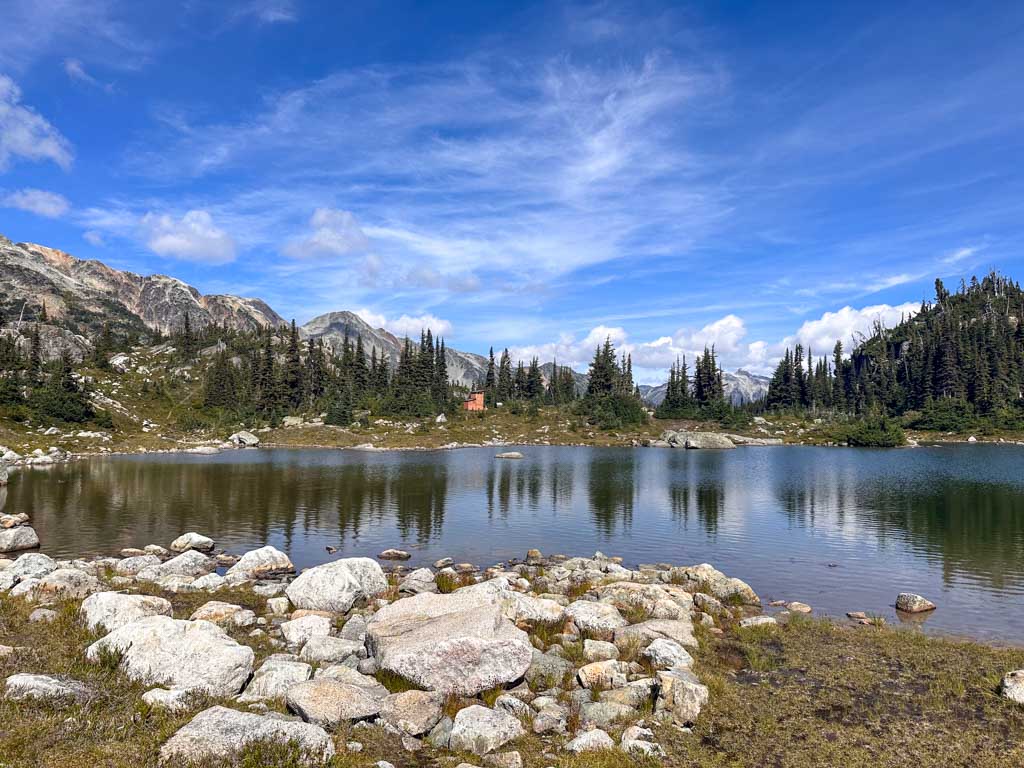 An alpine lake with rocks in the foreground. You can see a small wooden outhouse building on the other side of the lake. 