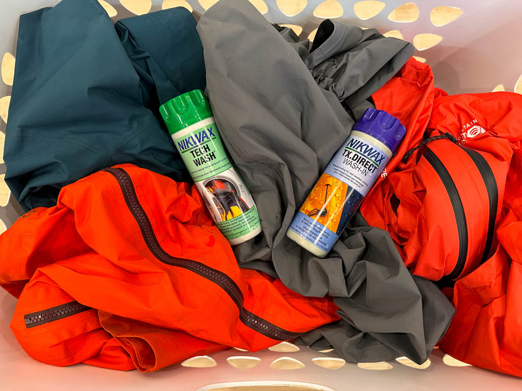 A basket of hiking rain jackets with bottles of tech wash and DWR retreatment. Why do rain jackets wet out? Because the DWR wears off.