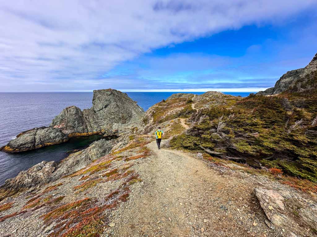 A man with a yellow backpack hikes along the winding Nanny Hole Trail towards a headland at Crow Head in Twillingate Newfoundland