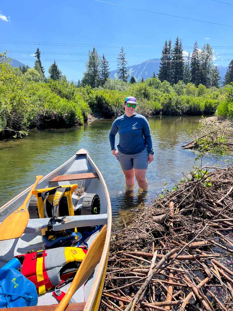 A woman stands in a river next to a beaver dam and a canoe. She is wearing the Mountain Hardwear Crater Lake Hoody