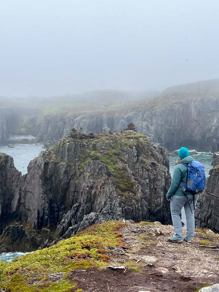 A woman stands at the edge of a cliff watching puffs on an offshore island near Bonavista. She is dressed for hiking with a backpack, warm jacket, and fleece hat.