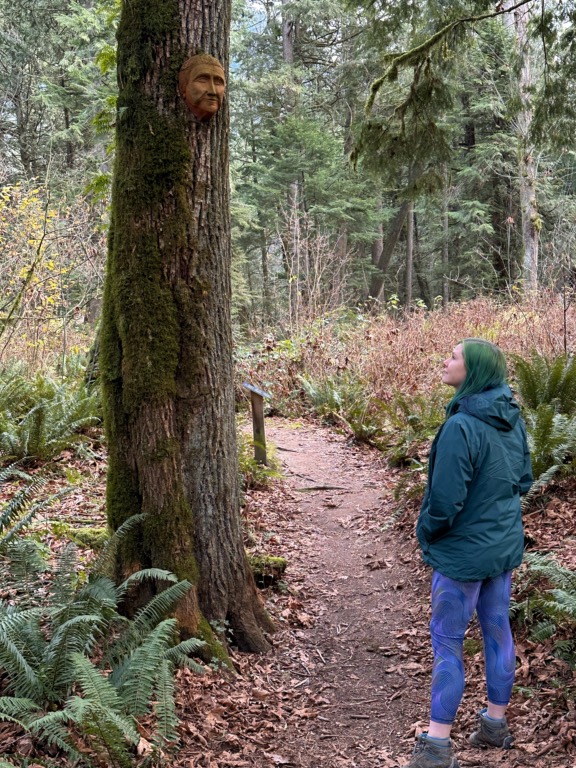 A woman in a green jacket looks at a mask mounted on a tree on the Spirit Trail