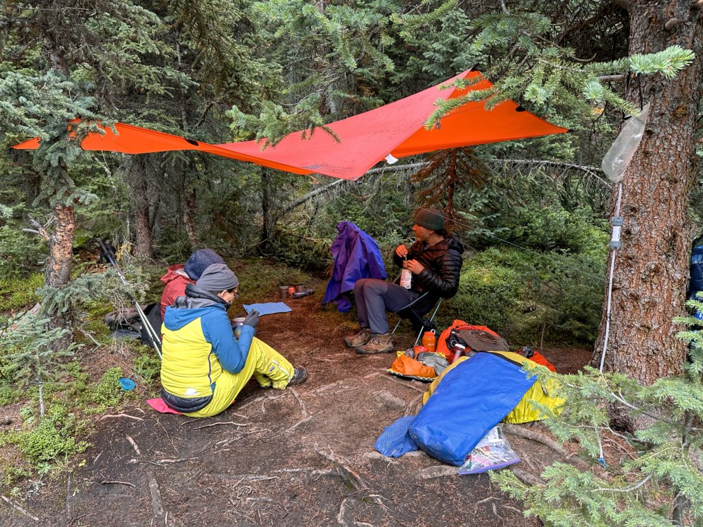 A group of hikers eats under an orange MEC Scout Silicone tarp - one of my picks for the best canadian hiking gear gifts