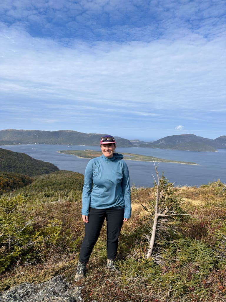 A woman in a long sleeved shirt and pants poses on a fall hike in Western Newfoundland