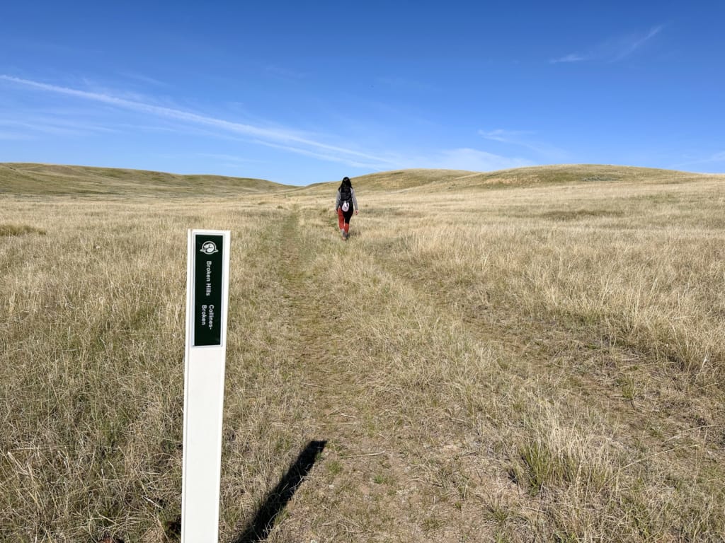 A woman walks past a trail marker in the grass on the Broken Hills Trail in Grasslands National Park