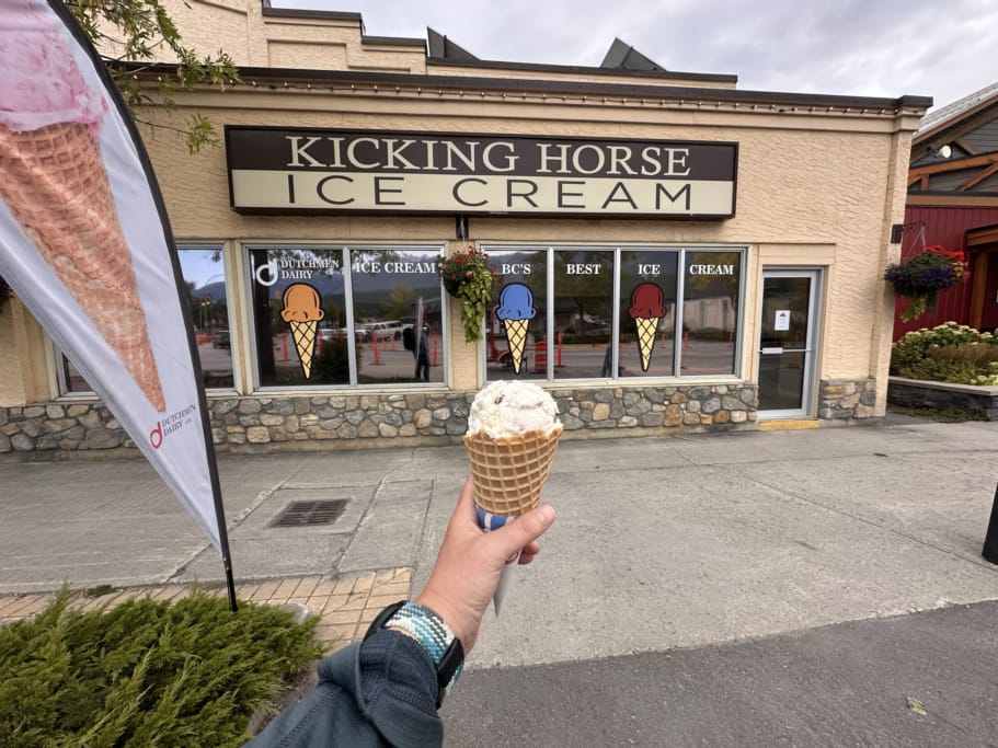 A person holds up an ice cream cone in front of the sign for Kicking Horse Ice Cream in Golden
